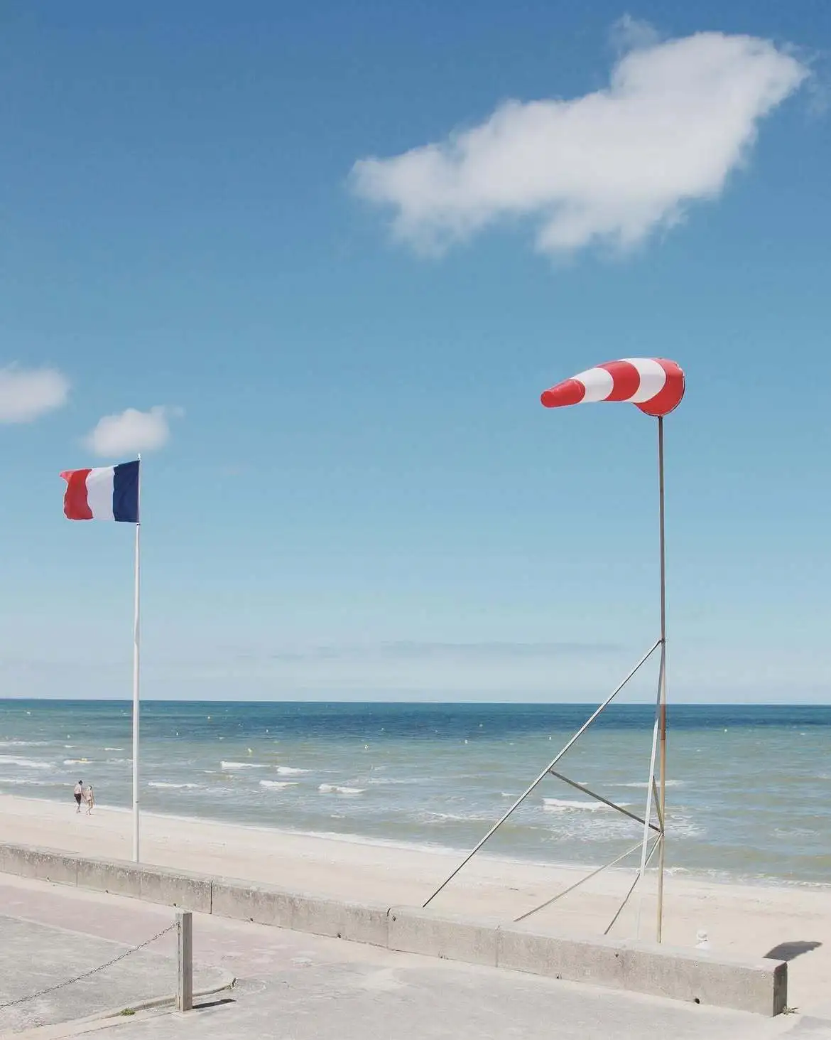 Une manche à air rouge et blanche et un drapeau français tendus par le vent au-dessus de la digue, la plage et la mer en contrebas.