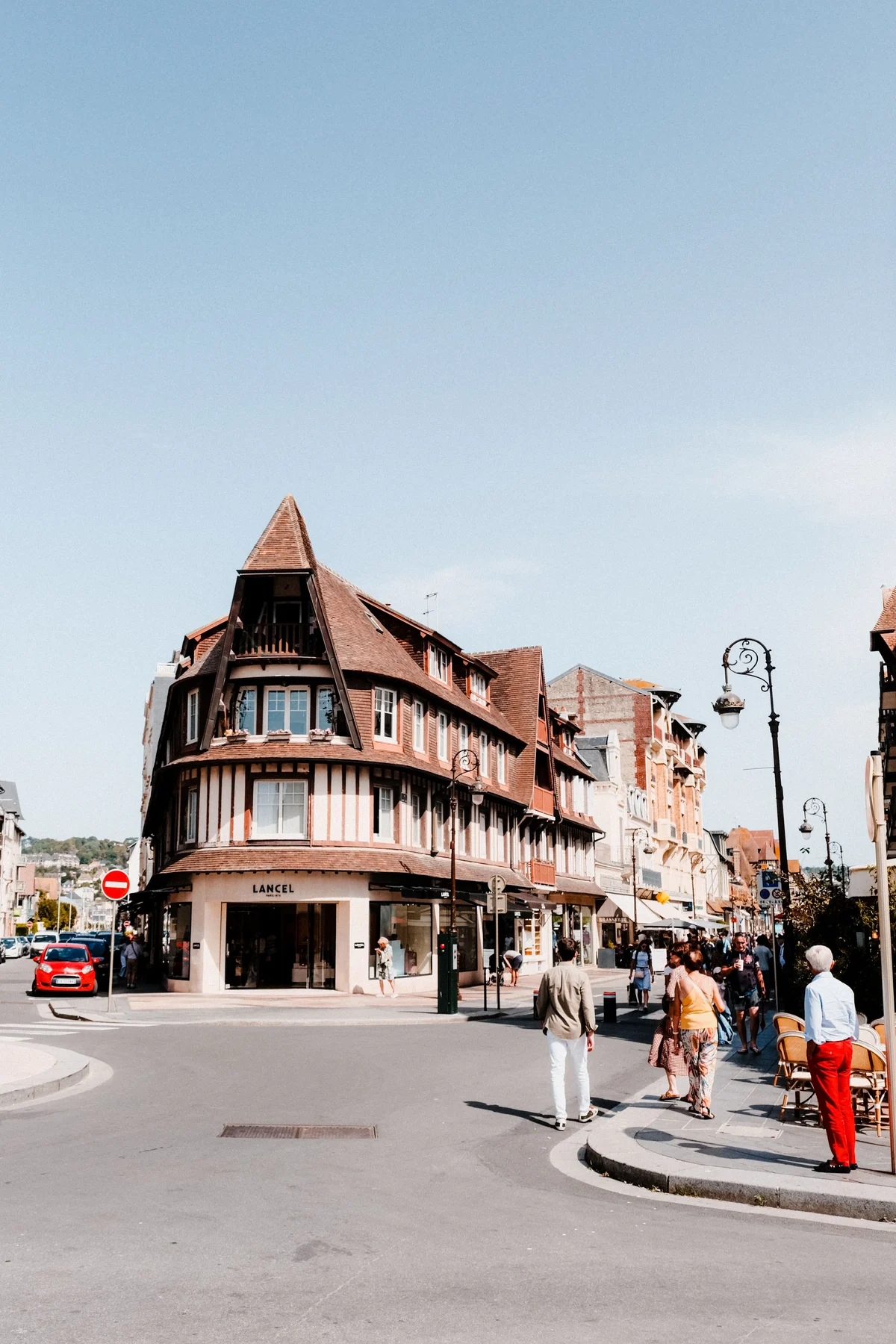 Un carrefour du centre de Deauville en plein soleil : immeuble d'angle à colombages coiffé d'une toiture pointue, boutiques en rez-de-chaussée et passants sur le trottoir.