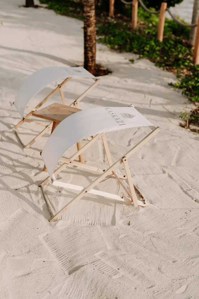 Deux tabourets pliants en bois et toile blanche posés sur le sable clair, à l'ombre d'un palmier.