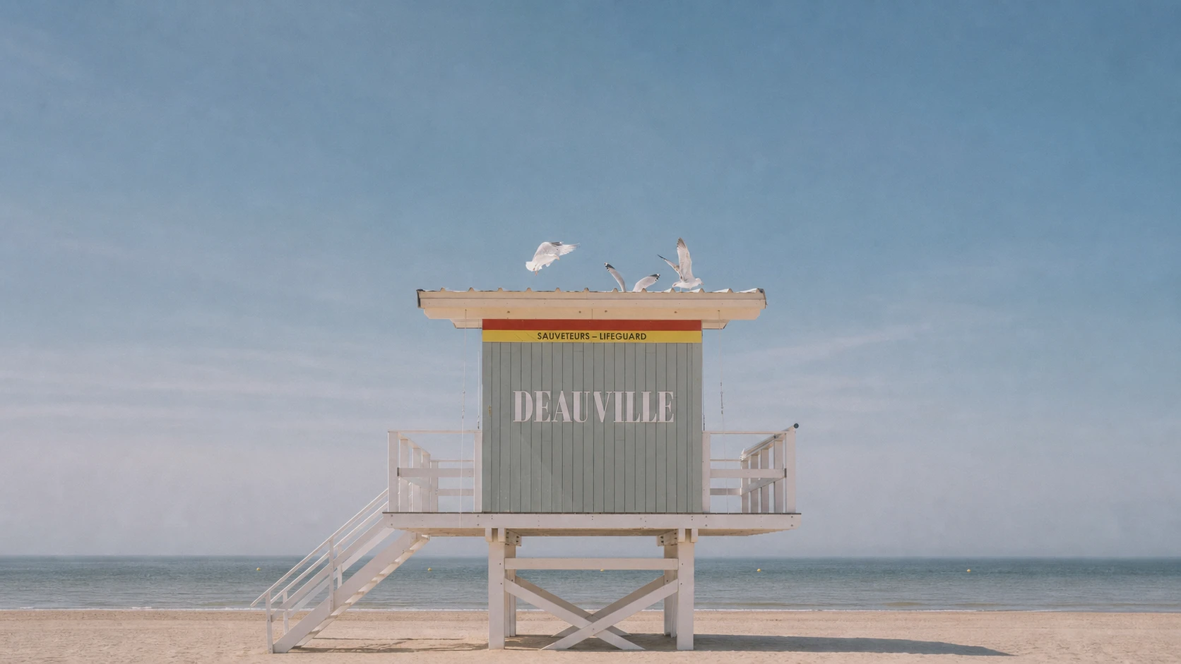 Poste de secours sur la plage de Deauville : une cabane gris-vert sur pilotis portant « DEAUVILLE » et « Sauveteurs / Lifeguard », des mouettes s'envolant du toit, la mer plate à l'horizon.
